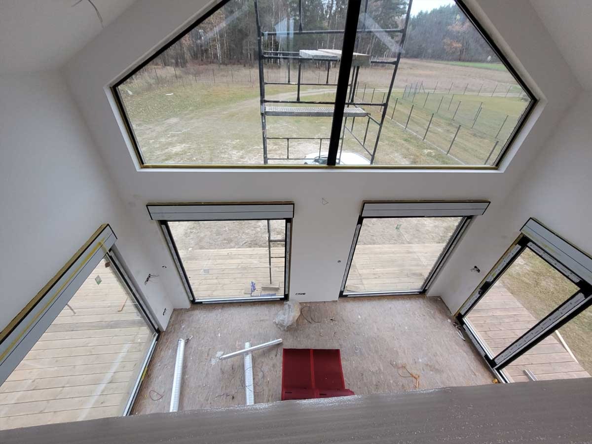 Wnętrze domu w trakcie wykańczania z dużym trójkątnym oknem dachowym i kilkoma mniejszymi oknami bocznymi, widok z góry na podłogę z widocznymi elementami konstrukcyjnymi i narzędziami.