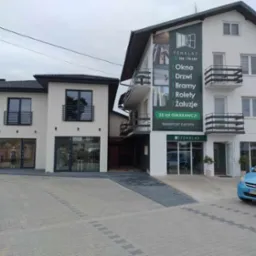 Elewacja budynku z reklamą firmy Fenklas oferującej okna, drzwi, bramy i rolety. Błękitny samochód zaparkowany przed budynkiem. Widoczny drugi budynek z czarnymi oknami.