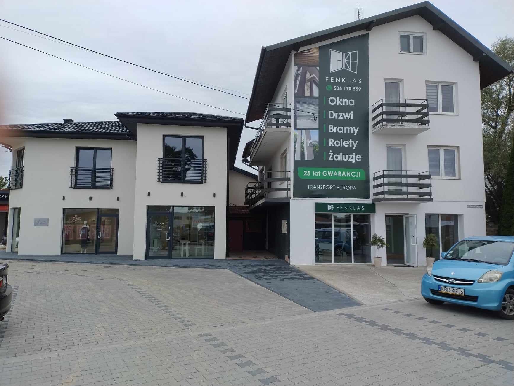 Elewacja budynku z reklamą firmy Fenklas oferującej okna, drzwi, bramy i rolety. Błękitny samochód zaparkowany przed budynkiem. Widoczny drugi budynek z czarnymi oknami.