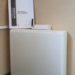 Centrala alarmowa Satel Integra z instrukcją obsługi i anteną, umieszczona w pomieszczeniu z widocznymi śladami użytkowania na ścianach i podłodze.