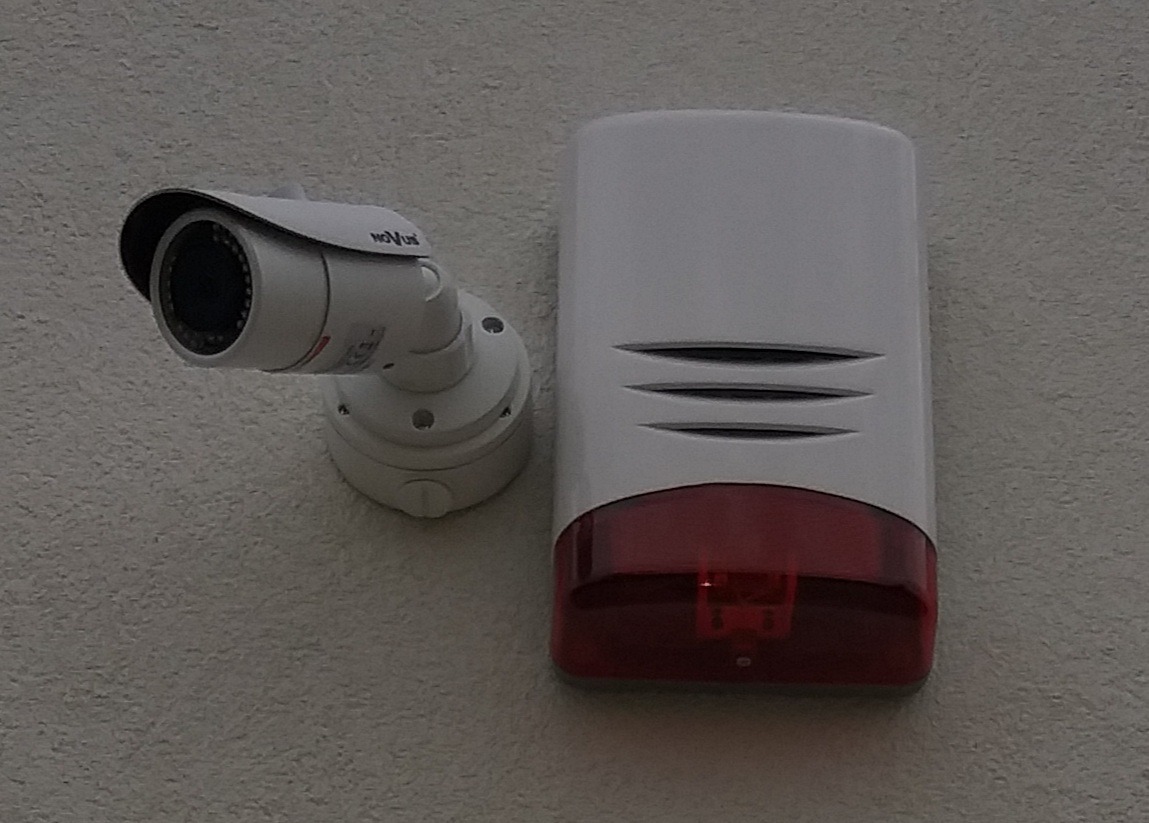 Kamera monitoringu marki Novus zamontowana obok sygnalizatora alarmowego z czerwoną szybką, na teksturowanej ścianie.