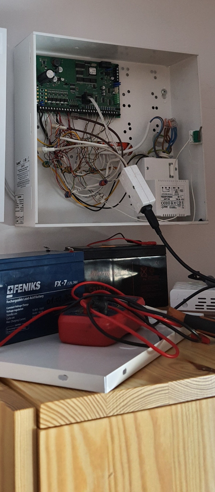 Otwarta obudowa centrali alarmowej z widoczną płytą główną, licznymi przewodami, transformatorem, akumulatorami FENIKS FX-7 i miernikiem elektrycznym na drewnianym blacie.