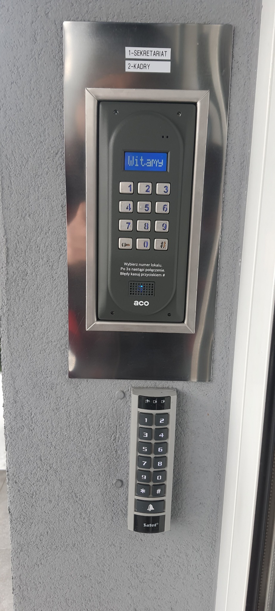 Dwa panele domofonowe z klawiaturą numeryczną, jeden marki ACO w metalowej ramce z wyświetlaczem LCD i napisem 'Witamy', drugi marki Satel, zamontowane na szarej, chropowatej ścianie przy wejściu...