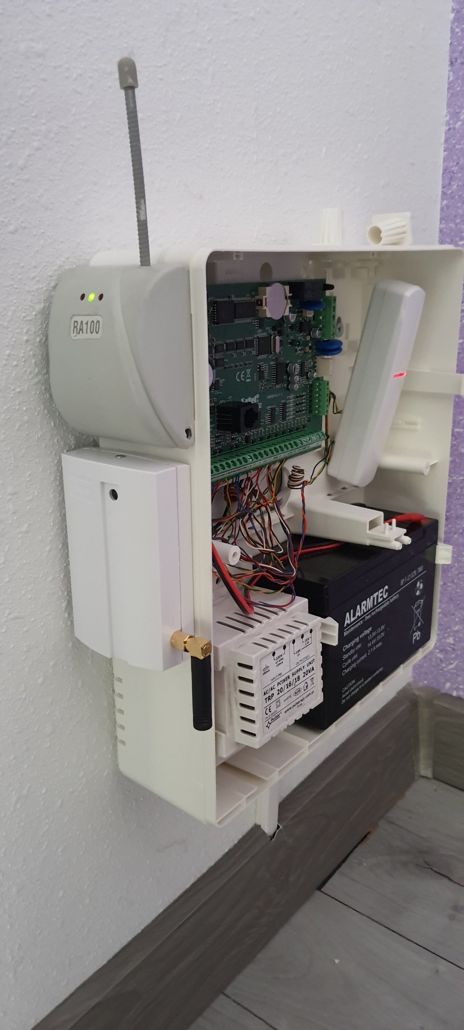 Otwarta obudowa centrali alarmowej RA100 z widoczną elektroniką, akumulatorem Alarmtec, zasilaczem Pulsar TRP 20/16/18 20VA i anteną, zamontowana na białej ścianie z szarymi panelami podłogowymi.
