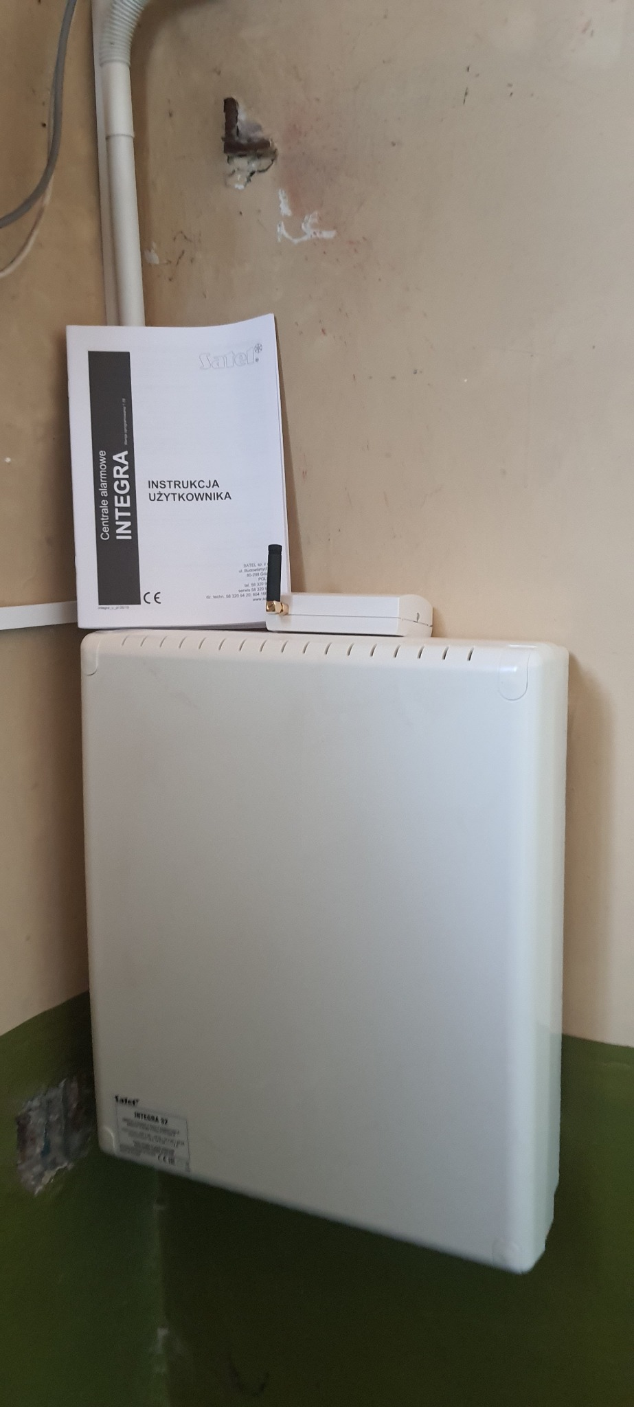Centrala alarmowa Satel Integra z instrukcją obsługi i anteną, umieszczona w pomieszczeniu z widocznymi śladami użytkowania na ścianach i podłodze.