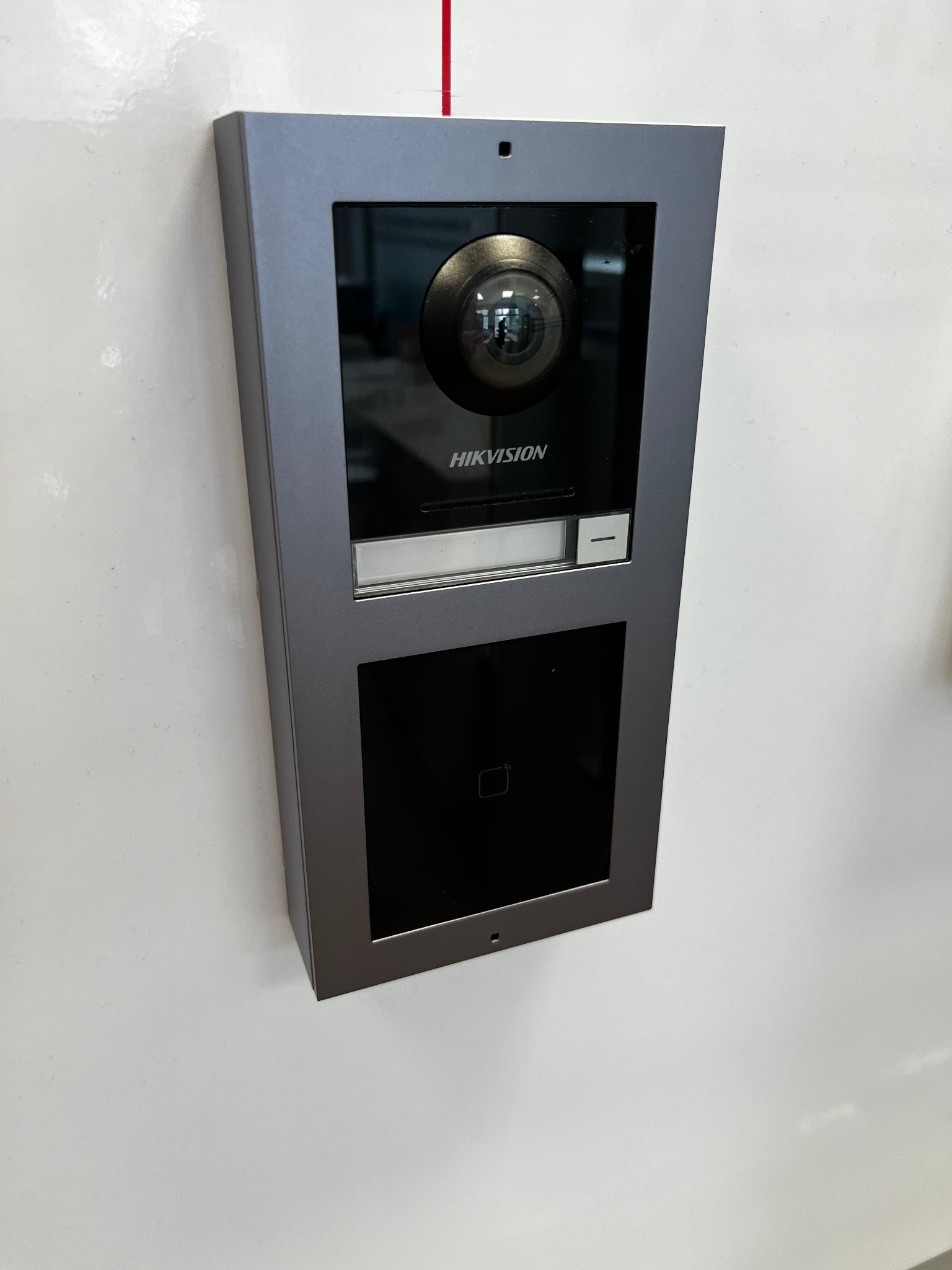 Szary wideofon marki Hikvision zamontowany na białej ścianie, widoczna kamera, czytnik kart i przycisk.