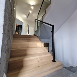 Usługi Stolarskie Szymon Ramus - Nowoczesne schody z drewna dębowego z czarną balustradą ze szkła, oświetlenie punktowe na suficie, widok z dołu na górę.