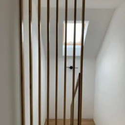 Nowoczesna, drewniana ażurowa balustrada schodowa z pionowych listew, jasne drewno, białe ściany, widok na schody i okno dachowe.