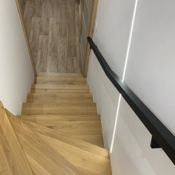 Usługi Stolarskie Szymon Ramus - Widok z góry na jasne, dębowe schody z czarną, minimalistyczną balustradą, oświetlone subtelnym światłem LED wzdłuż ściany, z fragmentem podłogi w tle.
