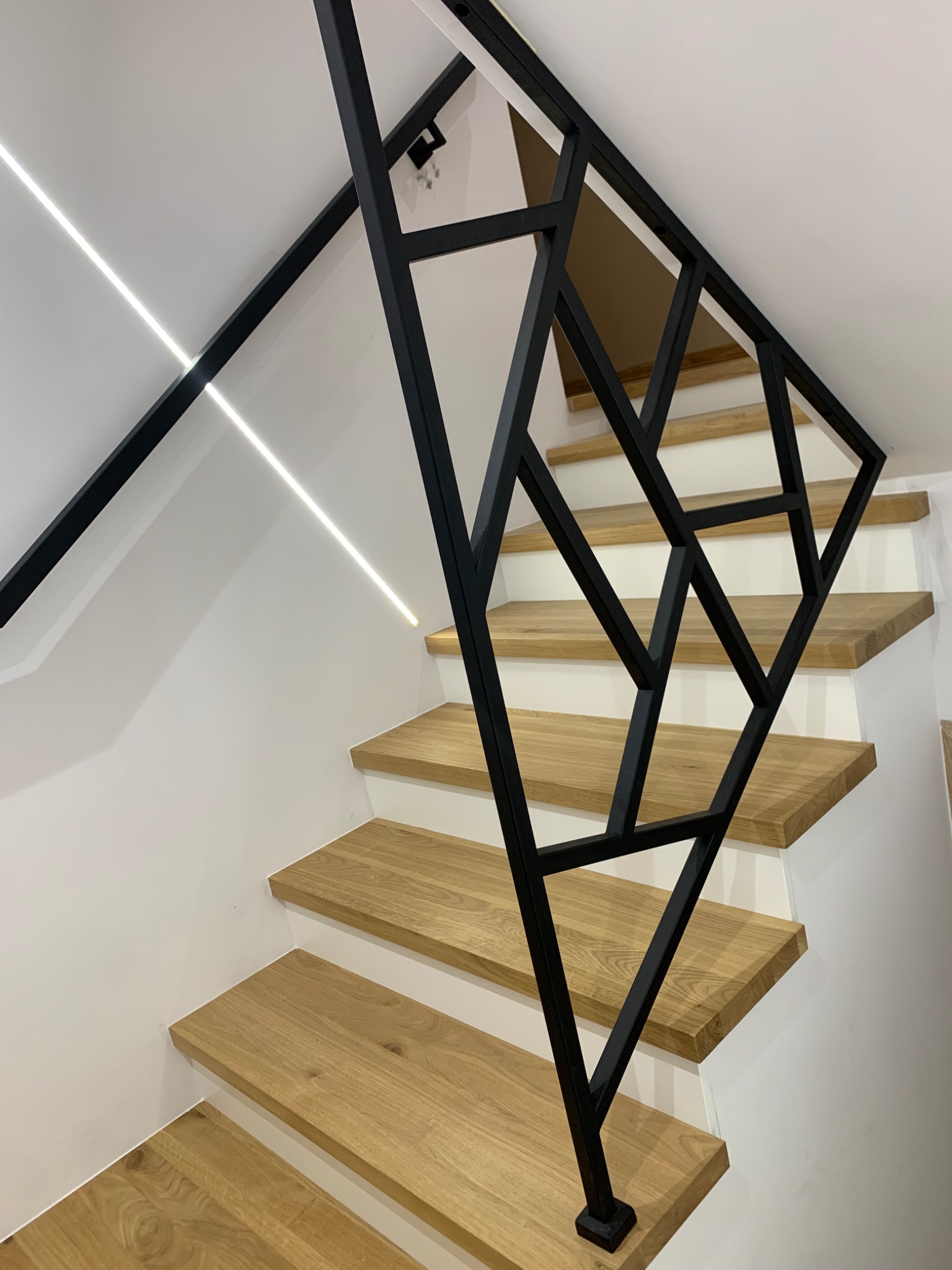 Nowoczesne schody z drewnianymi stopniami i czarną, geometryczną balustradą, oświetlone listwą LED wzdłuż ściany.