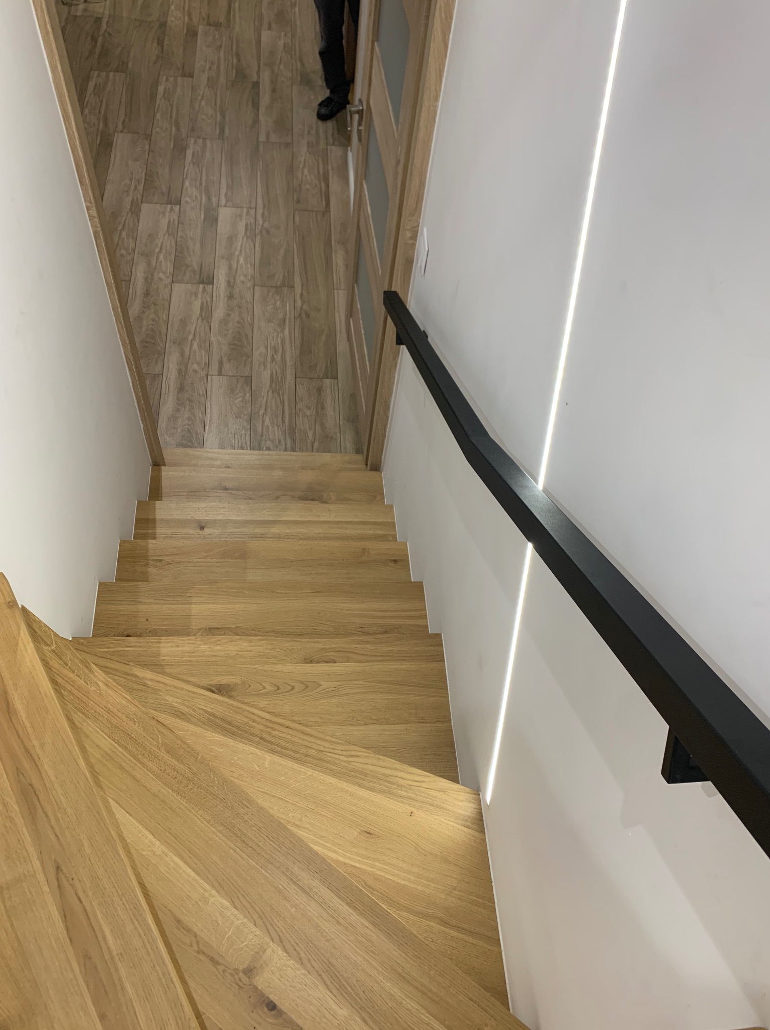Widok z góry na jasne, dębowe schody z czarną, minimalistyczną balustradą, oświetlone subtelnym światłem LED wzdłuż ściany, z fragmentem podłogi w tle.
