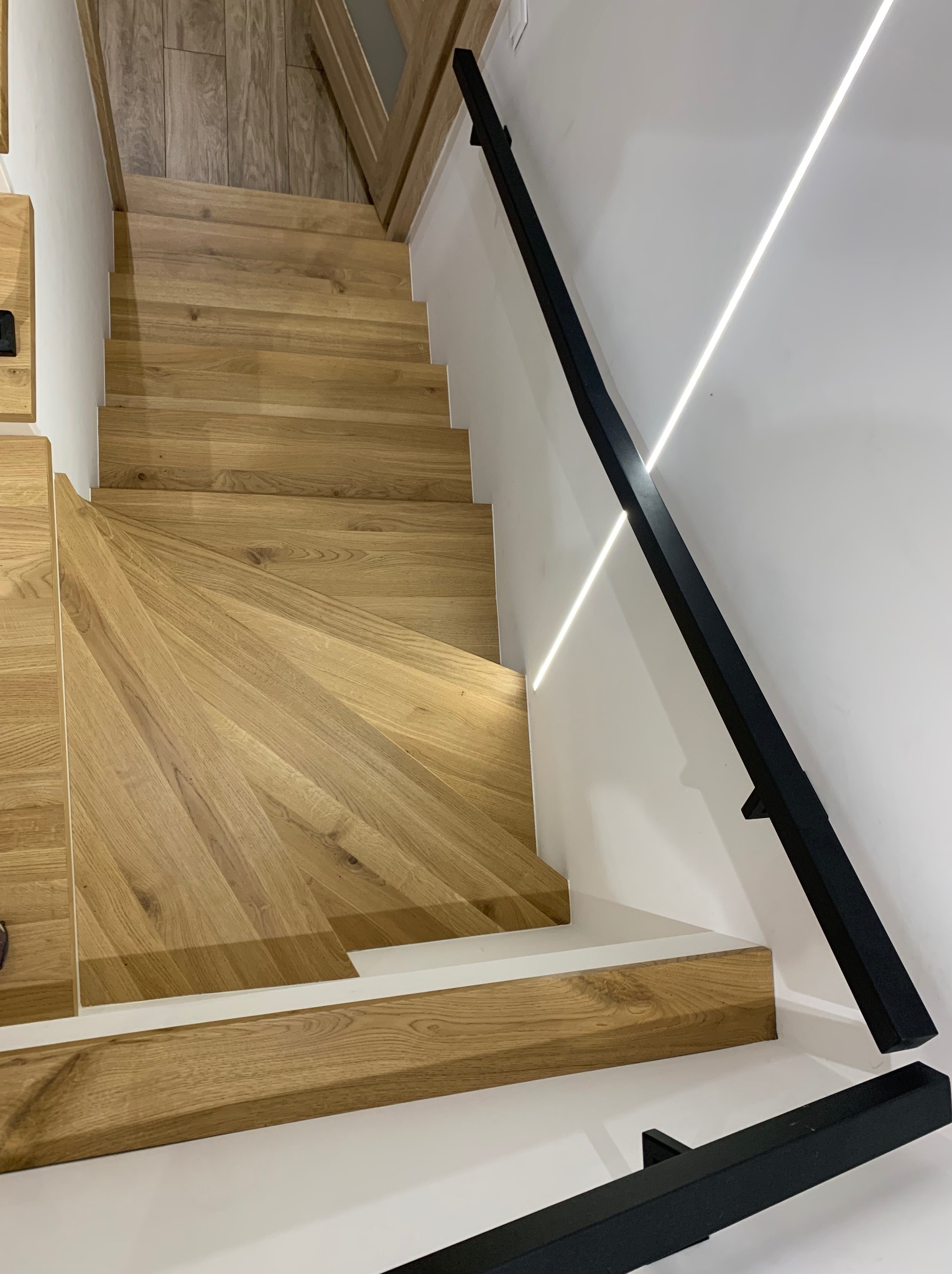 Widok z góry na jasne drewniane stopnie schodów z czarną, minimalistyczną balustradą na białej ścianie z oświetleniem LED.