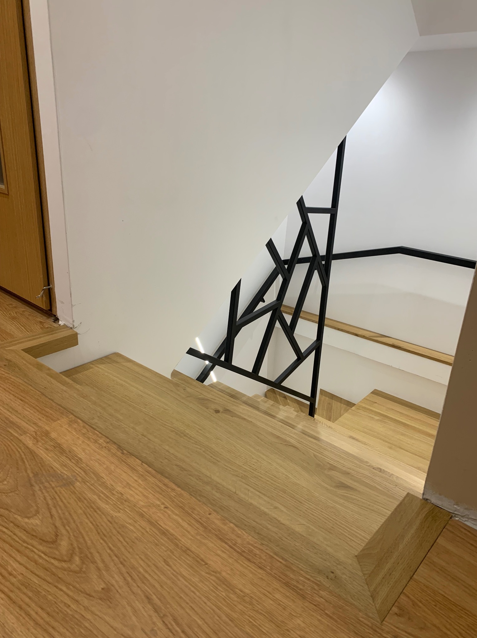 Widok z góry na drewniane schody z nowoczesną, geometryczną balustradą z czarnego metalu w jasnym wnętrzu.