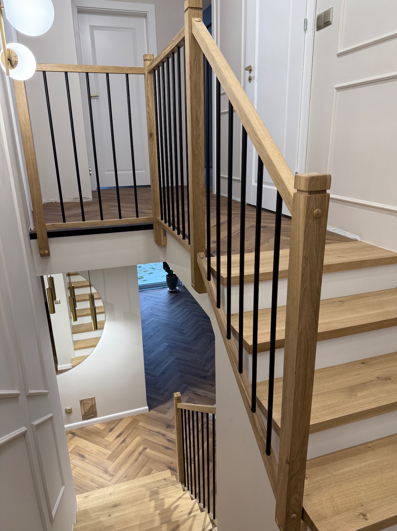 Widok z góry na drewniane schody z jasnego drewna z czarnymi, metalowymi balustradami, widoczne również drewniane podłogi ułożone w jodełkę oraz lustro odbijające schody.