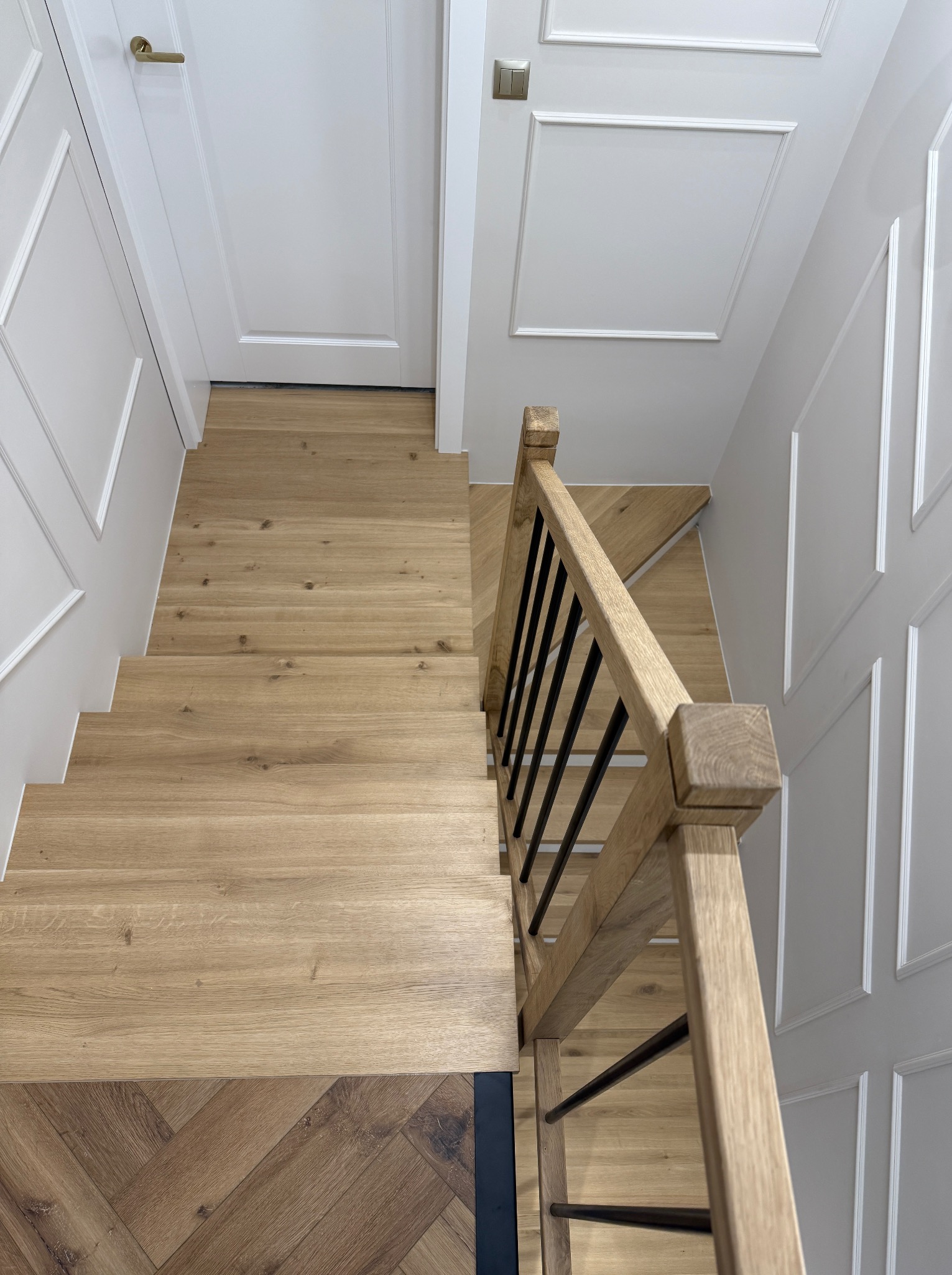 Widok z góry na jasne, drewniane schody z balustradą z naturalnego drewna i czarnymi prętami, kontrastujące z ciemniejszą, ułożoną w jodełkę podłogą na dole, białe ściany z dekoracyjnymi listwami.