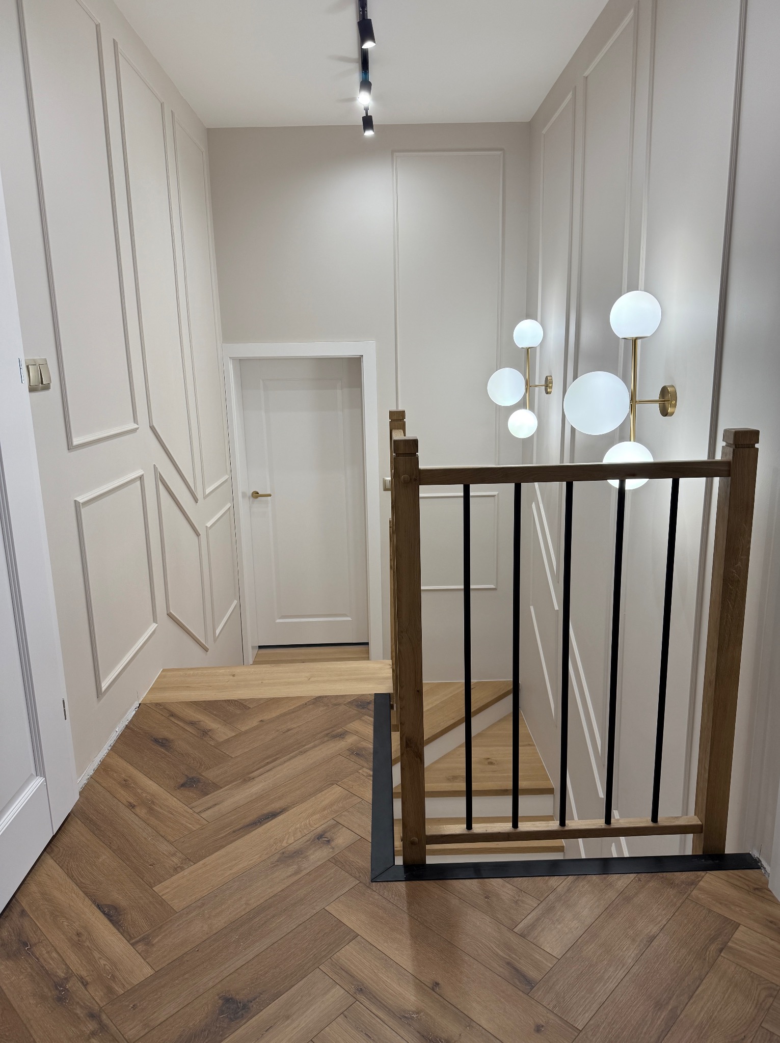 Nowoczesne wnętrze z drewnianymi schodami, balustradą z czarnymi elementami i podłogą ułożoną w jodełkę, oświetlone lampami kulami na złotych ramionach, z widocznym fragmentem białych drzwi...
