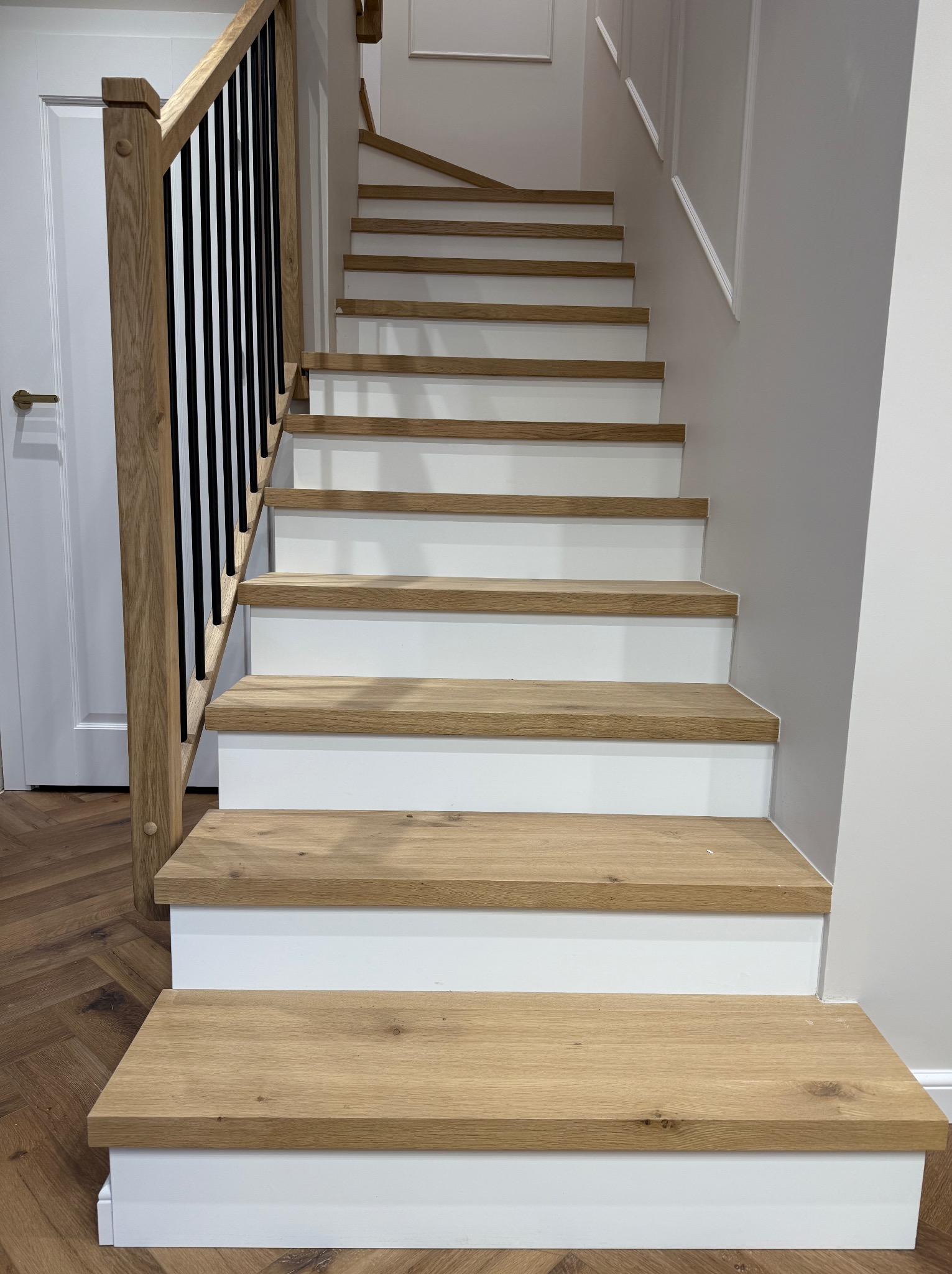 Nowoczesne schody z drewnianymi stopniami i białymi podstopnicami, z czarną balustradą zamontowaną na drewnianym słupku, widok od dołu na tle parkietu ułożonego w jodełkę.