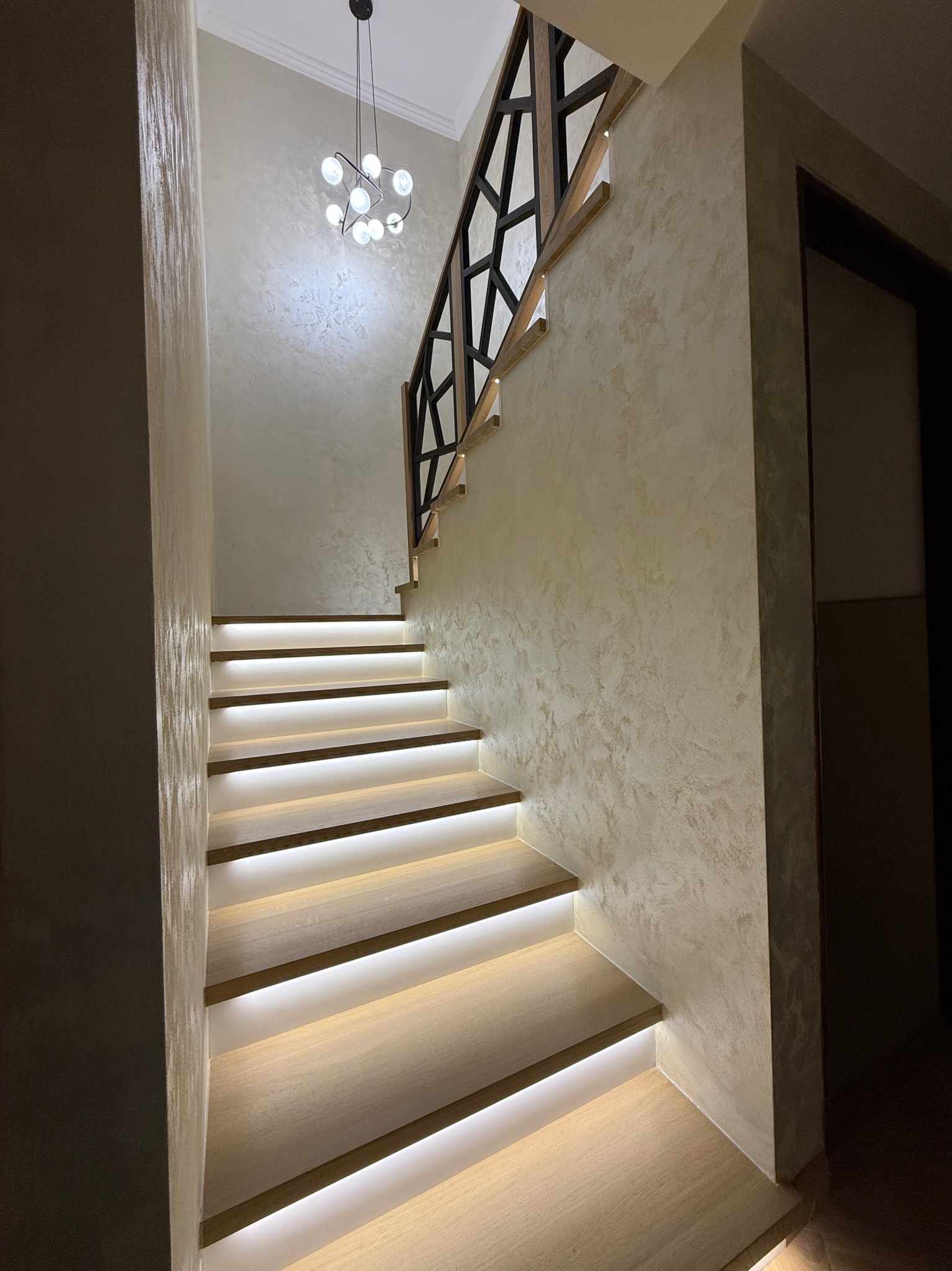 Nowoczesne schody z podświetleniem LED i balustradą o geometrycznym wzorze. Ściany wykończone dekoracyjnym tynkiem. Ujęcie od dołu, widoczny fragment sufitu z lampą.