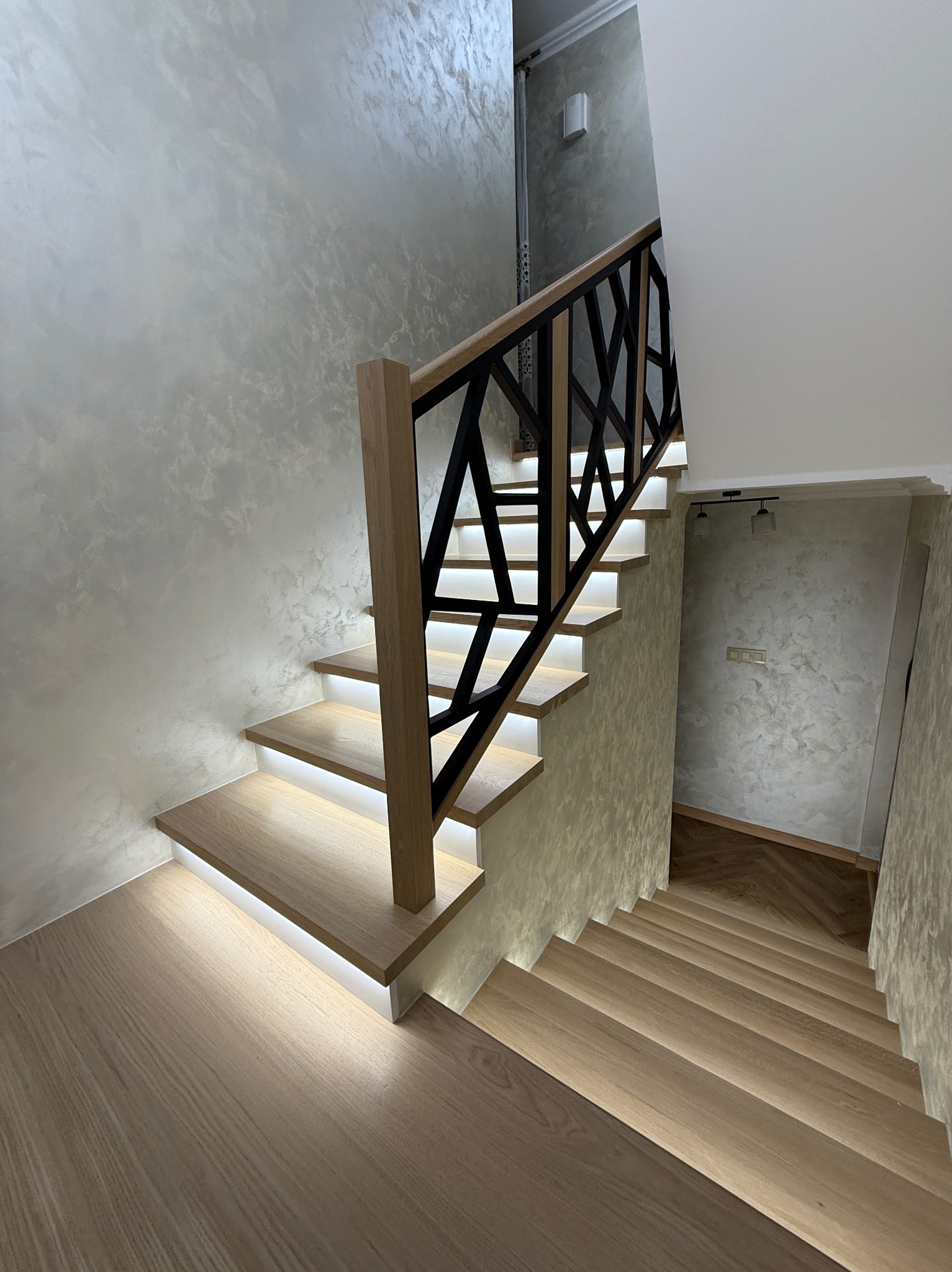 Nowoczesne schody z podświetleniem LED i geometryczną, czarną balustradą na tle ściany o fakturze betonu. Widok z góry.