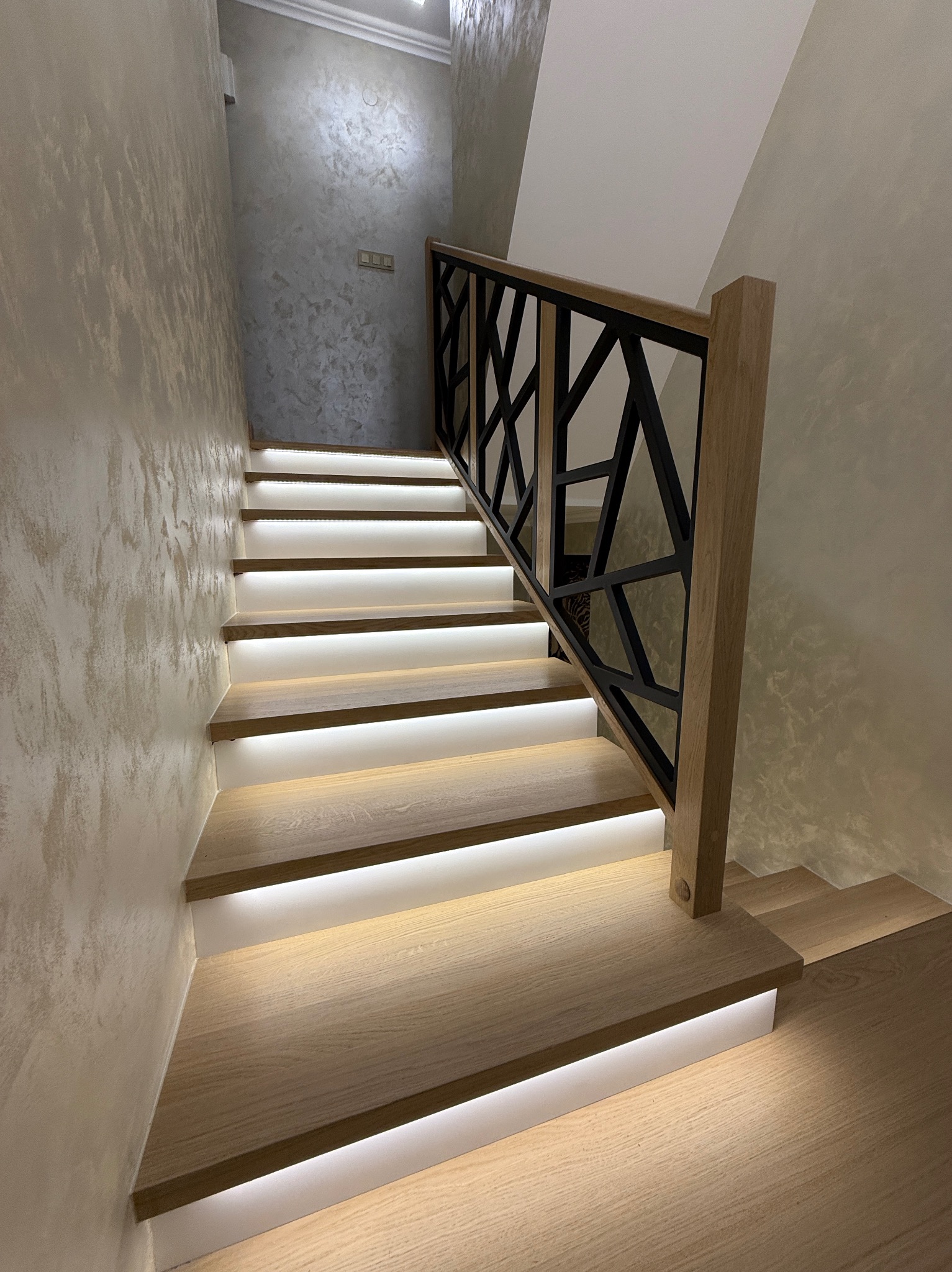 Nowoczesne schody z podświetleniem LED i geometryczną, czarną balustradą. Jasne drewno stopni kontrastuje z ciemnym wzorem balustrady.