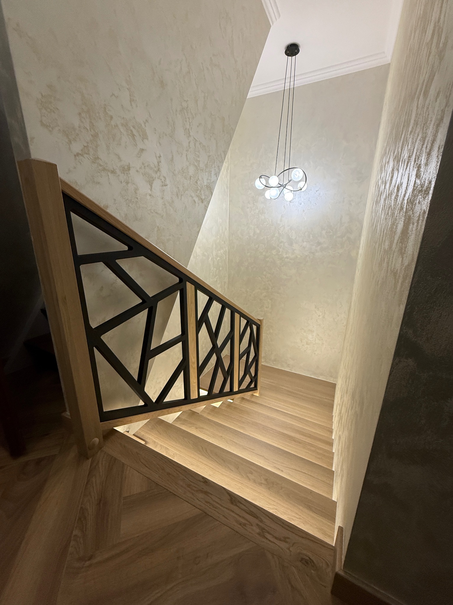 Nowoczesne schody z jasnego drewna z geometryczną, czarną balustradą. Parkiet w jodełkę i designerska lampa wisząca dopełniają elegancki wystrój wnętrza.