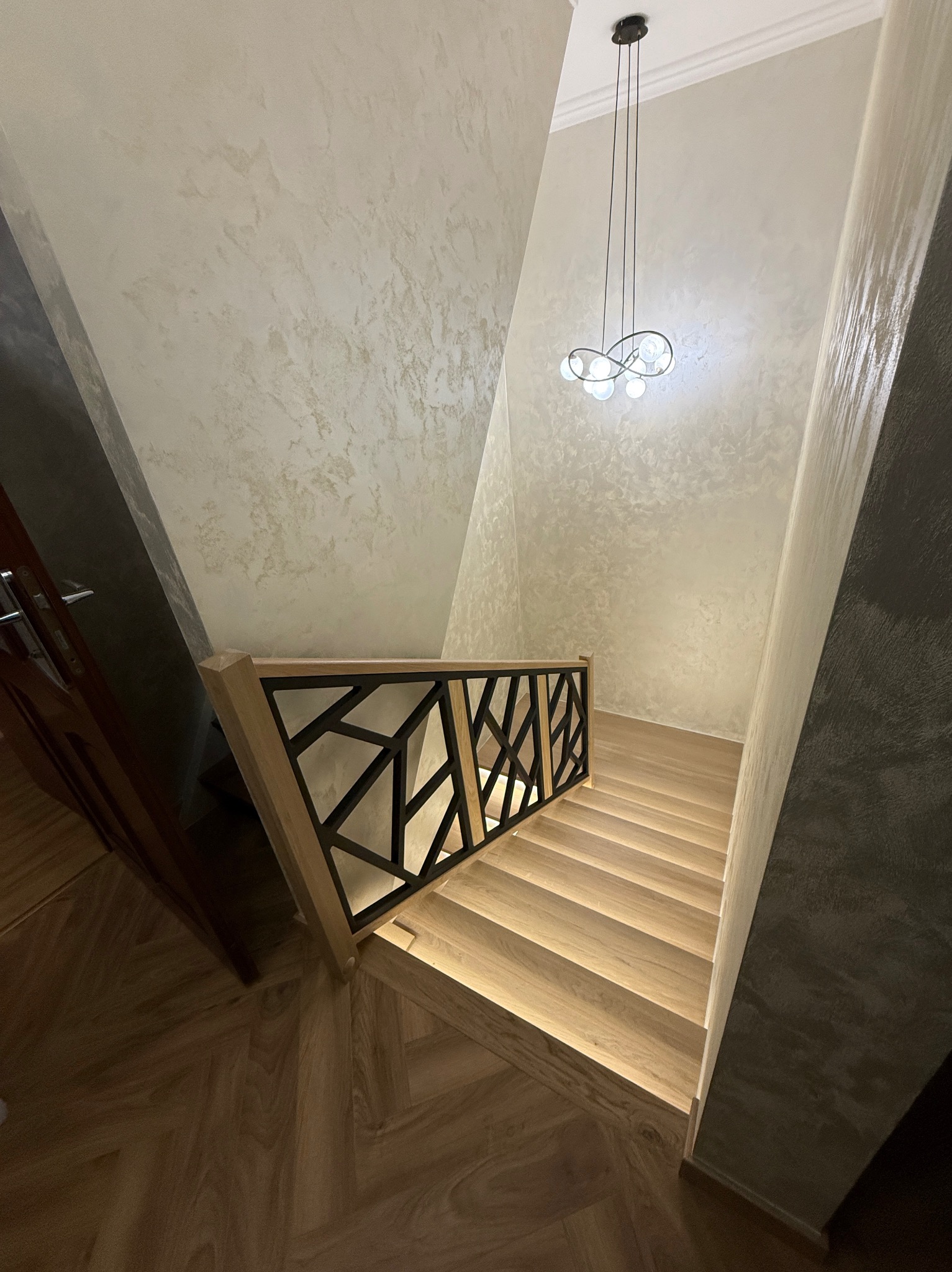 Nowoczesne schody z jasnego drewna z geometryczną balustradą i podświetleniem stopni, widok z góry, dekoracyjna lampa wisząca, ściany z efektem stiuku.