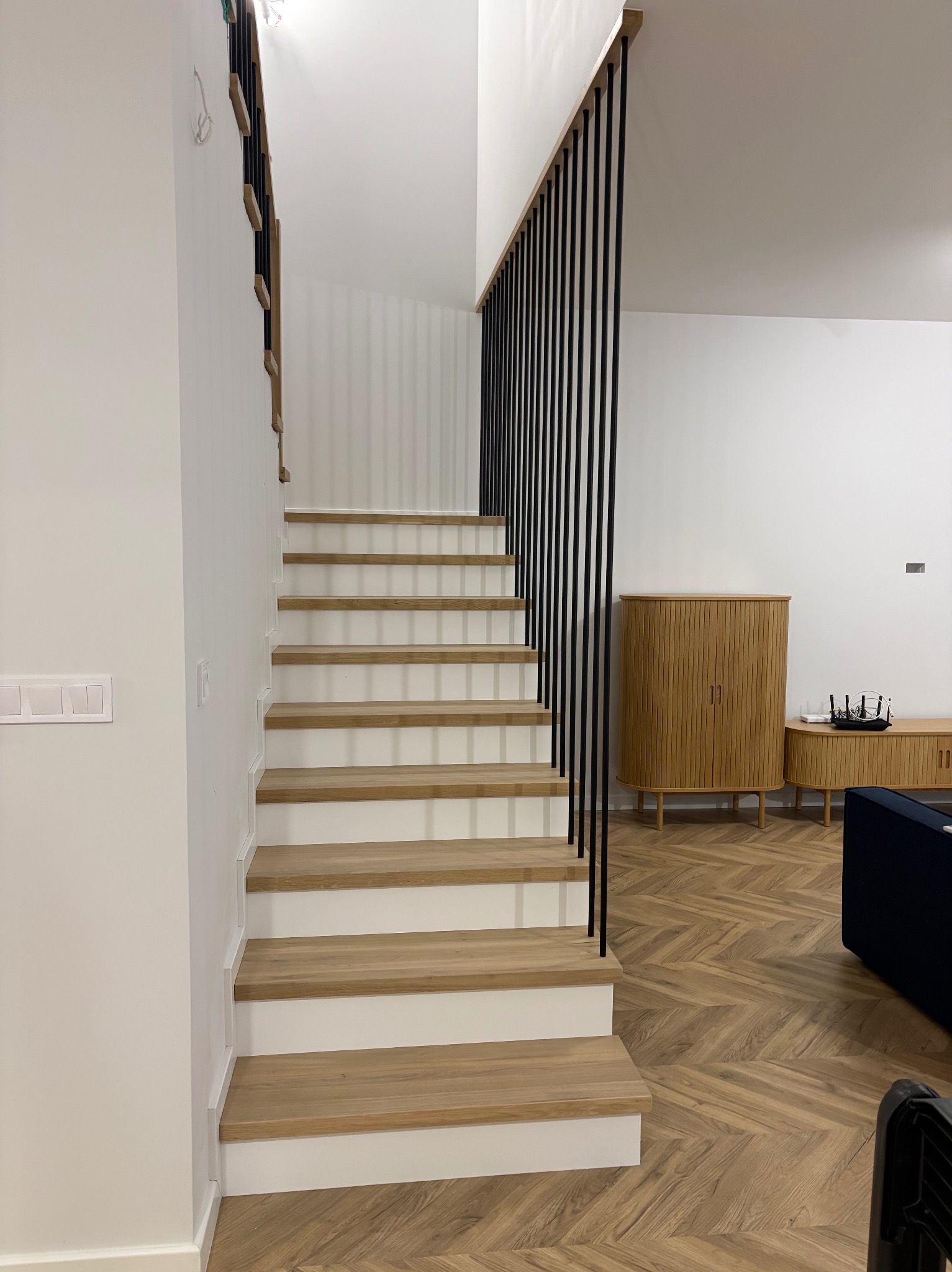 Nowoczesne schody z drewnianymi stopniami i minimalistyczną, czarną balustradą w jasnym wnętrzu. Parkiet w jodełkę i drewniana komoda w tle.
