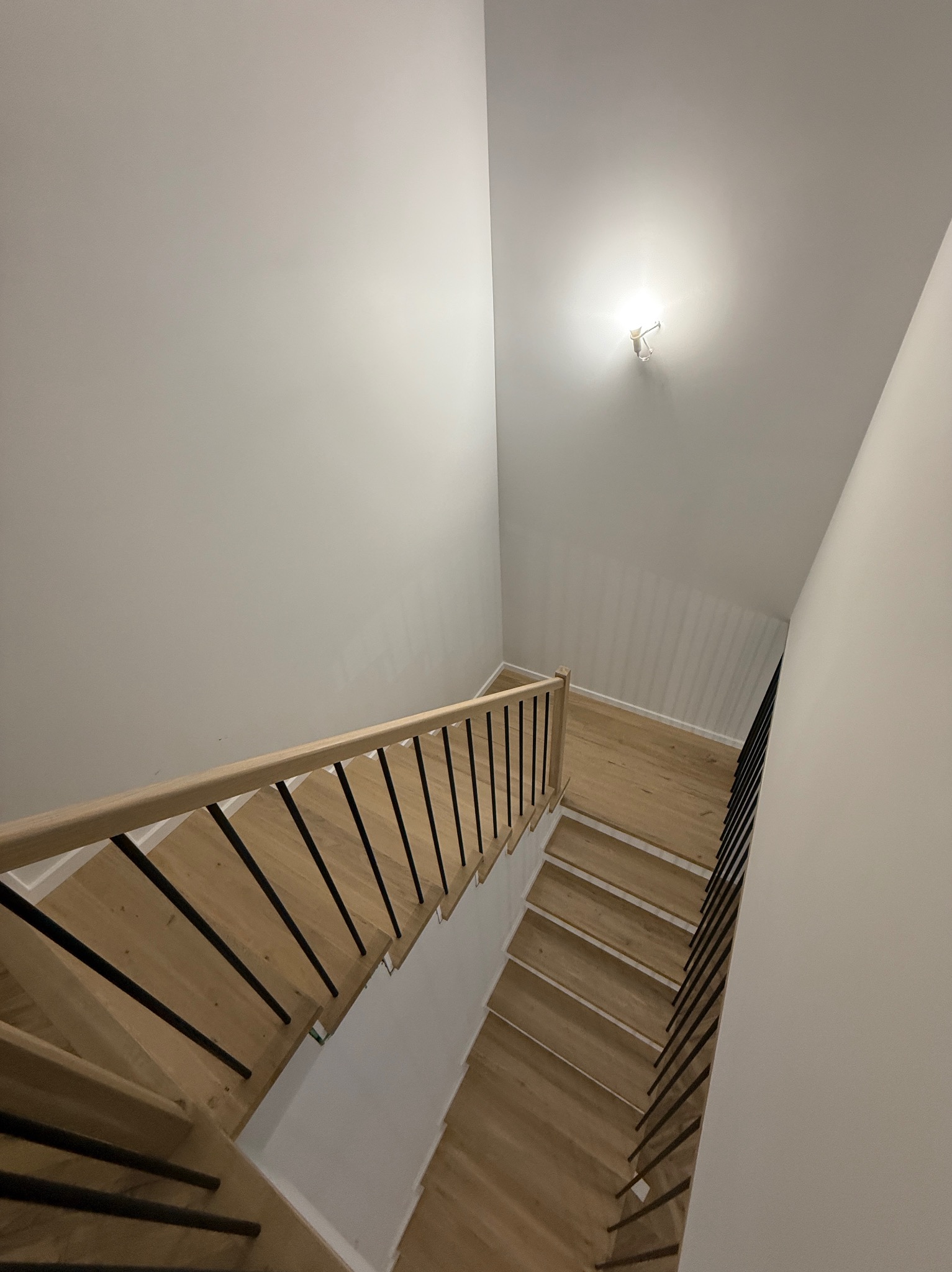 Jasne, drewniane schody z czarną, metalową balustradą, widziane z góry, oświetlone punktowym światłem na białym suficie. Minimalistyczny design.