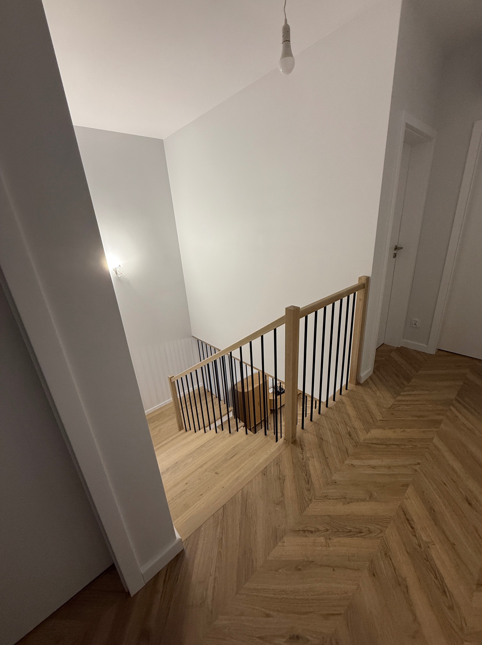 Jasne schody z drewnianymi stopniami i czarnymi, pionowymi balustradami, widziane z góry, z parkietem ułożonym w jodełkę i minimalistycznym oświetleniem.