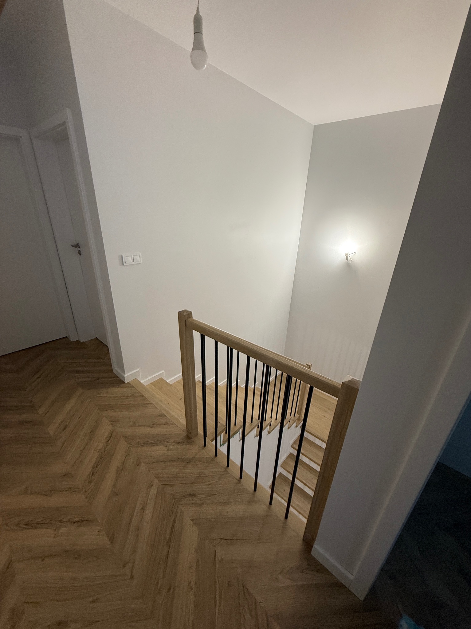 Nowoczesne schody z drewnianymi stopniami i czarnymi balustradami, widok z góry, parkiet w jodełkę na podłodze, minimalistyczne oświetlenie.