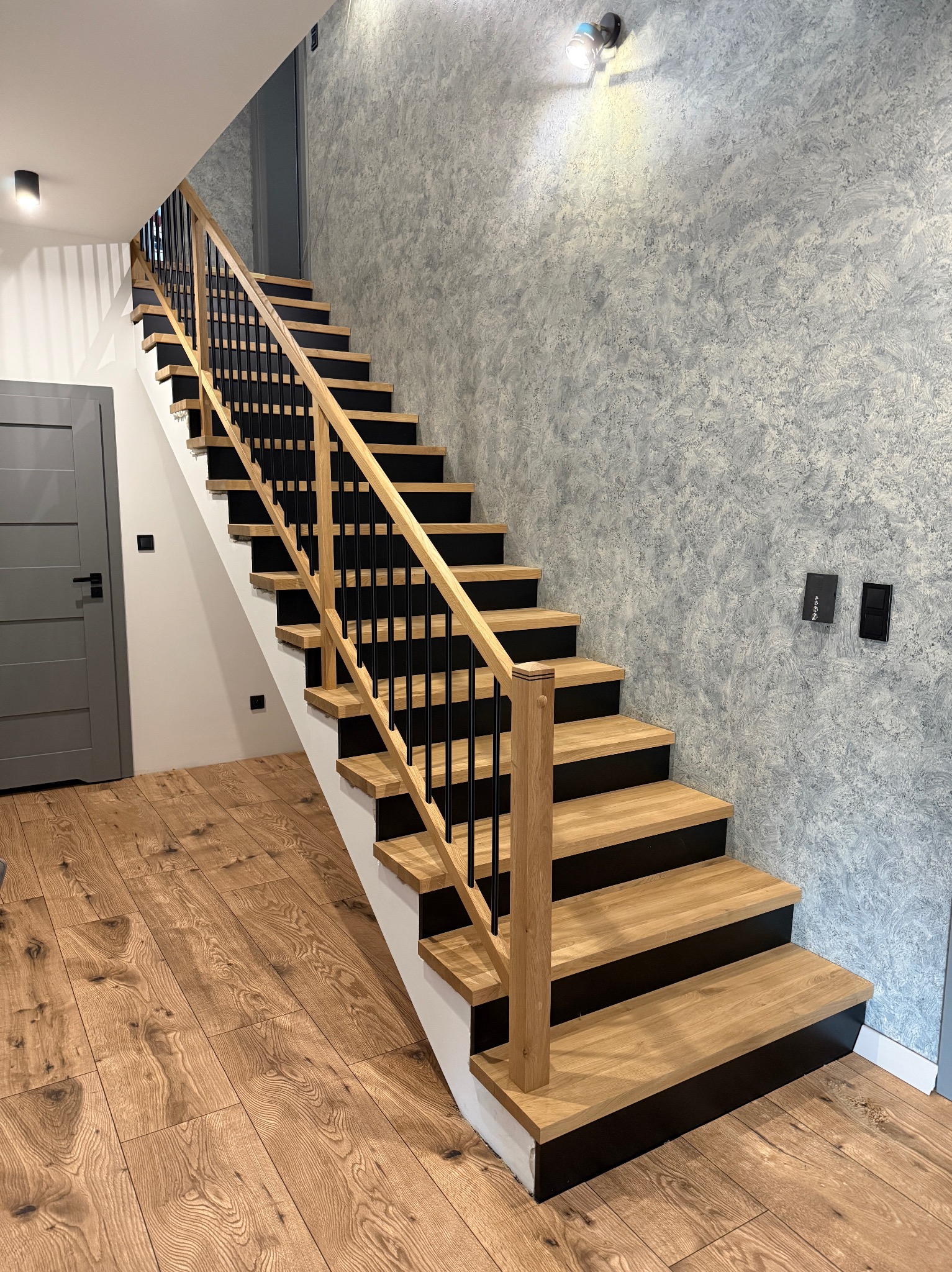Nowoczesne schody z drewnianymi stopniami i czarnymi podstopnicami, balustrada z jasnego drewna z czarnymi elementami, na tle ściany o fakturze betonu.