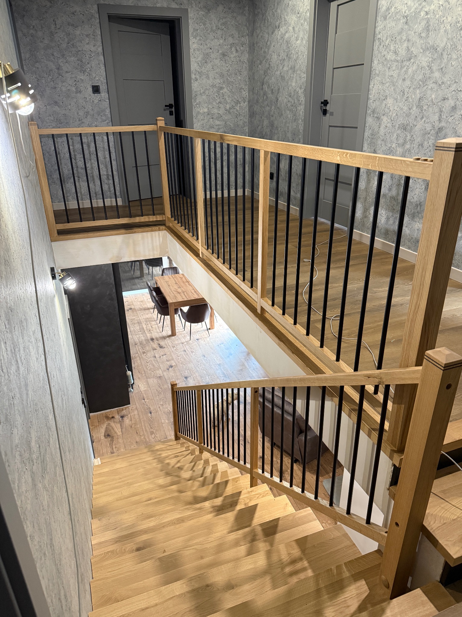 Drewniane schody z balustradą z czarnymi szczebelkami, widok z góry. Jasne drewno schodów i balustrady kontrastuje z szarymi ścianami i drzwiami w tle.
