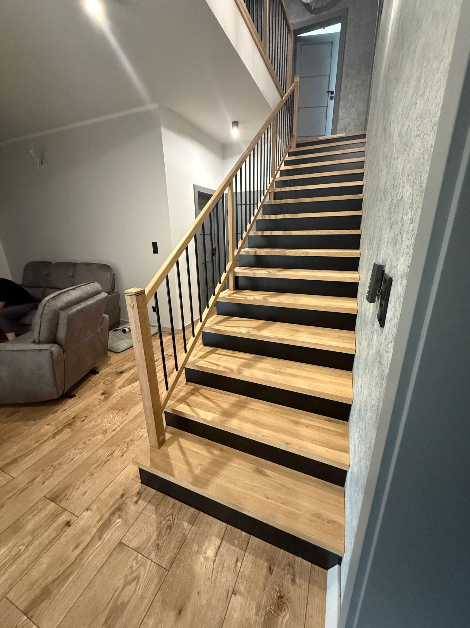 Nowoczesne schody z drewnianymi stopniami i czarną balustradą w jasnym wnętrzu. Widoczne drewniane podłogi i szara ściana z włącznikiem.