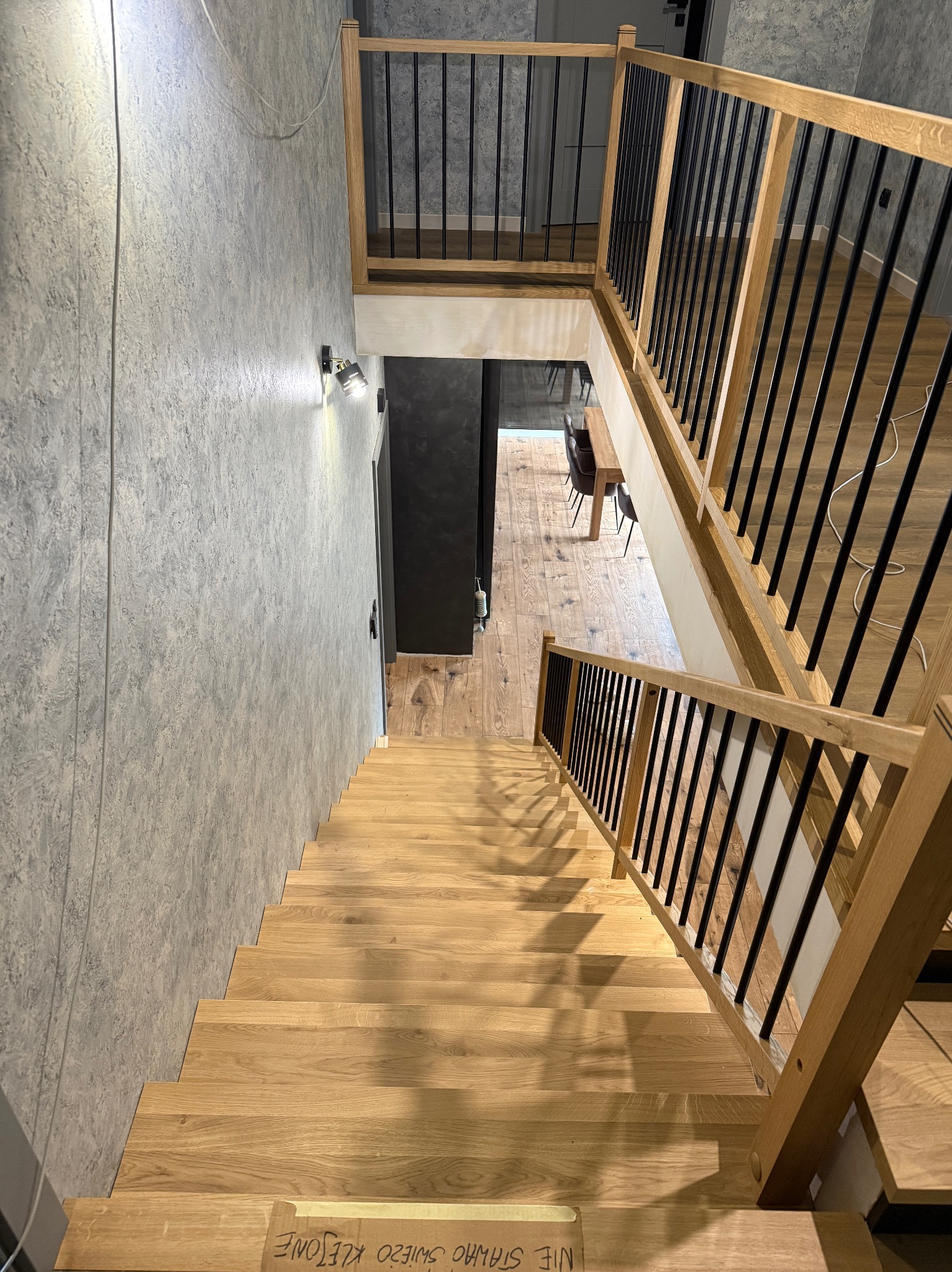 Nowoczesne schody z jasnego drewna z czarnymi balustradami, widok z góry. Na dole schodów karton z napisem. Minimalistyczny design, szara ściana w tle.