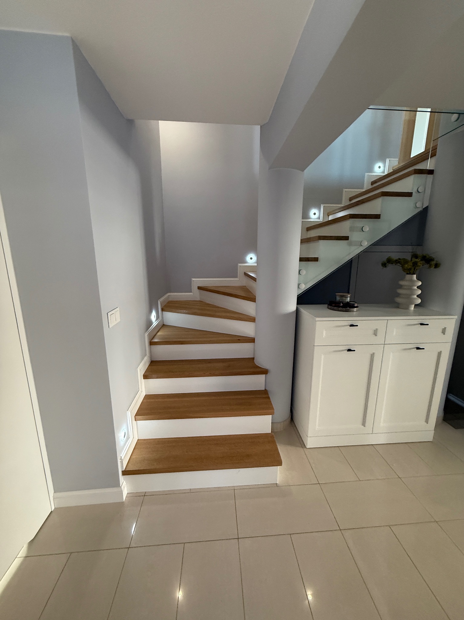 Nowoczesne schody z drewnianymi stopniami i szklaną balustradą, oświetlone punktowo, z białą szafką u dołu. Jasne wnętrze, minimalistyczny design.