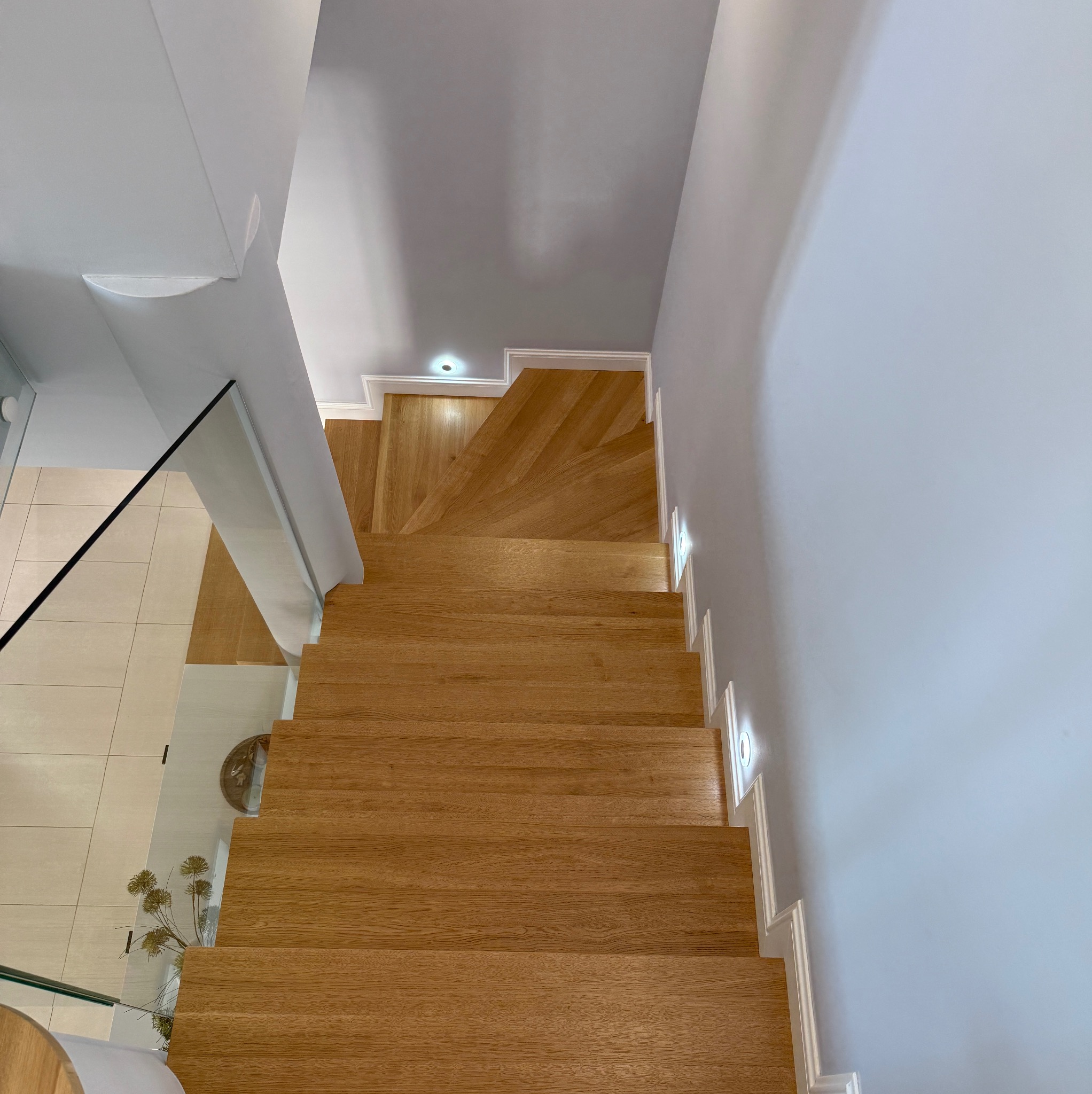 Widok z góry na jasne, dębowe schody z oświetleniem punktowym wzdłuż ściany i szklaną balustradą. Minimalistyczny design z elementami dekoracyjnymi.