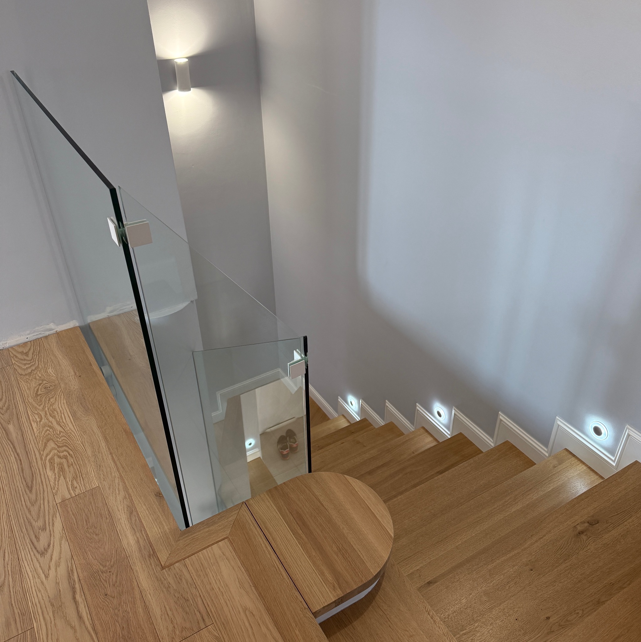 Nowoczesne schody z jasnego drewna z szklaną balustradą, oświetlone punktowymi lampami w ścianie. Widok z góry, ukazujący detale wykończenia i minimalistyczny design.