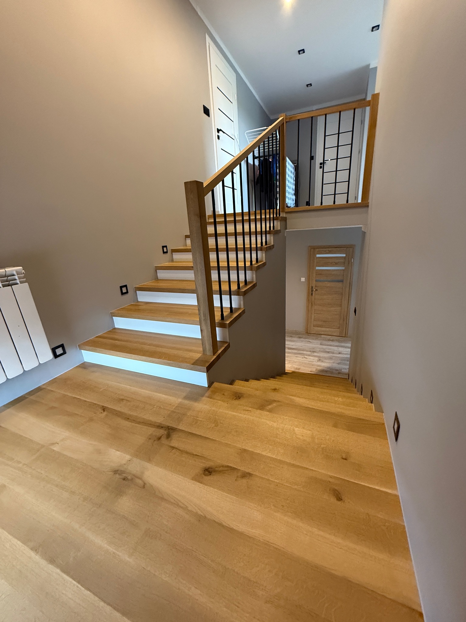 Drewniane schody z czarną balustradą i białymi podstopnicami w nowoczesnym wnętrzu z oświetleniem punktowym. Widok z dołu na górę.