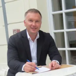 Mężczyzna w garniturze podpisuje dokument na okrągłym, białym stoliku na tle jasnej ściany i okna z kratownicą.