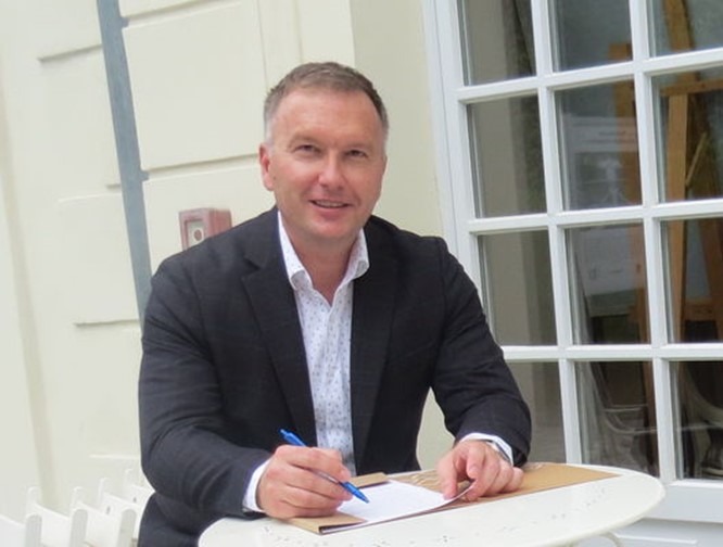 Mężczyzna w garniturze podpisuje dokument na okrągłym, białym stoliku na tle jasnej ściany i okna z kratownicą.