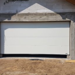JANDA OKNA - Biała segmentowa brama garażowa zamontowana w surowym stanie budynku z betonu komórkowego i widocznymi przewodami elektrycznymi.