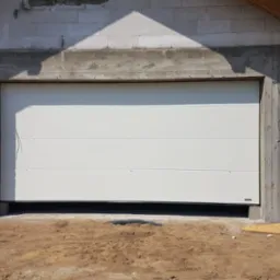 Biała segmentowa brama garażowa zamontowana w surowym stanie budynku z betonu komórkowego i widocznymi przewodami elektrycznymi.