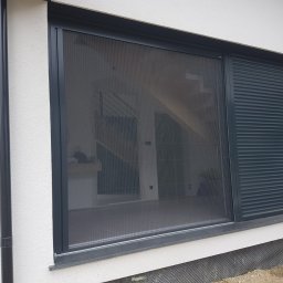 JANDA OKNA - Nowoczesne okno z moskitierą plisowaną i częściowo zasłoniętą roletą zewnętrzną w kolorze antracytowym, widok z zewnątrz na tle białej elewacji domu.