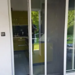 Przesuwne drzwi tarasowe z moskitierą, widok z zewnątrz na taras z desek kompozytowych, w tle wnętrze kuchni z żółtymi frontami.