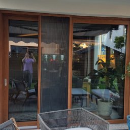 JANDA OKNA - Drewniane drzwi przesuwne z moskitierą plisowaną, widok z zewnątrz na taras z meblami ogrodowymi i odbiciem osoby robiącej zdjęcie.