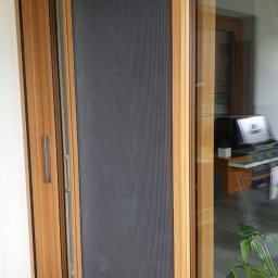 JANDA OKNA - Przesuwne okno PCV z szarą roletą plisowaną w drewnopodobnej ramie, widok z zewnątrz na taras.