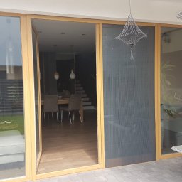 JANDA OKNA - Duże przesuwne drzwi tarasowe z moskitierą i drewnianą ramą, widok na wnętrze domu z jadalnią i schodami, odbicie osoby robiącej zdjęcie.