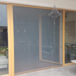 JANDA OKNA - Duże przesuwne okno z moskitierą plisowaną, widok z zewnątrz na taras, odbicie osoby robiącej zdjęcie, wiszące ozdobne lampy.