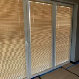 Trzyczęściowe białe okno z beżowymi żaluzjami poziomymi, widoczne zabezpieczenie podłogi folią malarską.