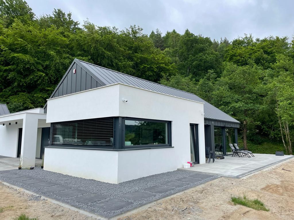 Nowoczesny dom jednorodzinny z białymi ścianami i grafitowymi oknami, z widokiem na zalesione wzgórze w tle, podkreślony przez minimalistyczną architekturę i starannie ułożony chodnik z szarych płyt.